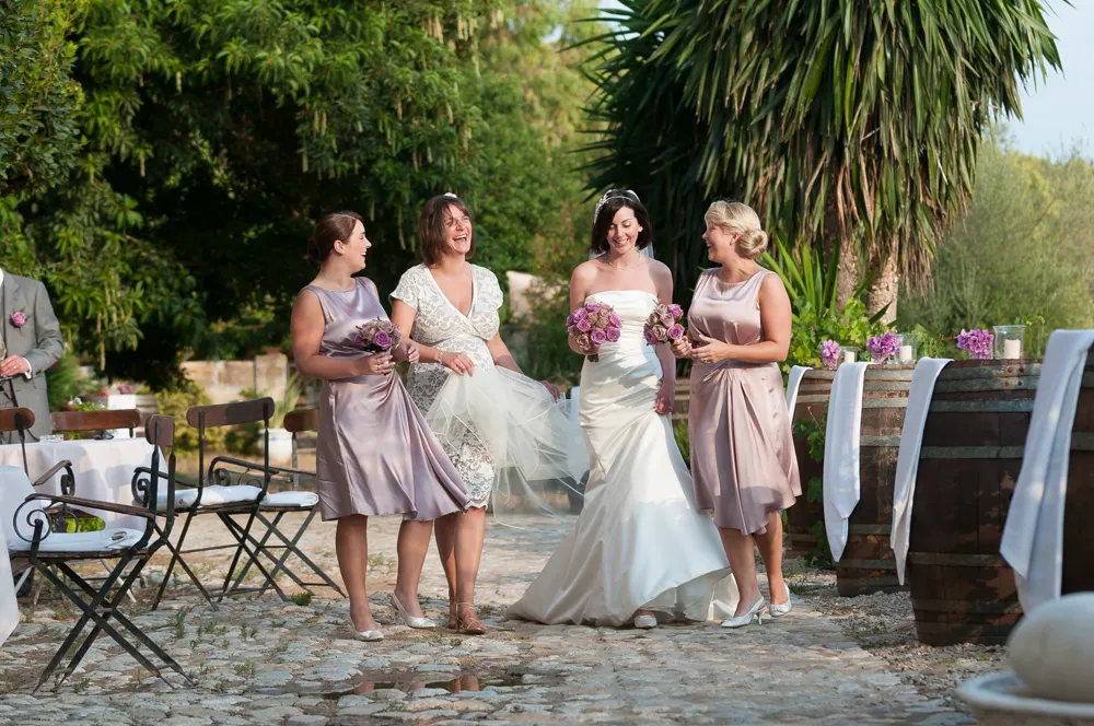 Bride in white gown holding purple bouquets walking with three bridesmaids in lavender dresses on a cobblestone path outdoors.