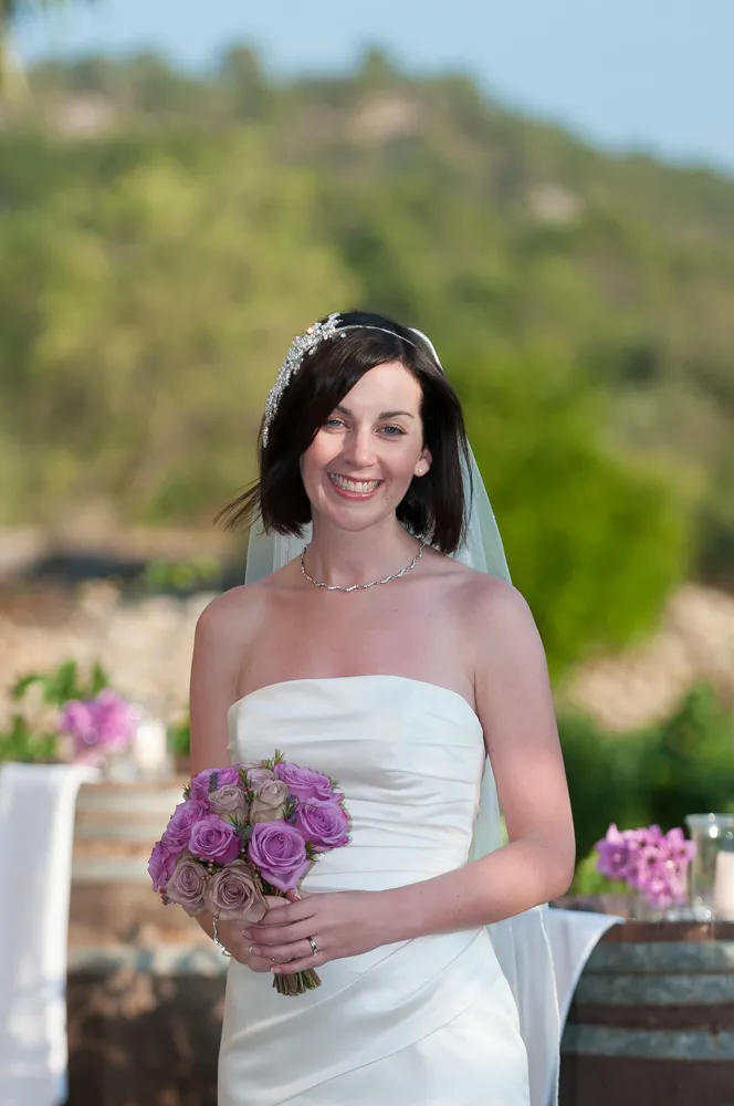 Smiling bride in strapless white wedding dress holding a bouquet of purple and beige roses outdoors.