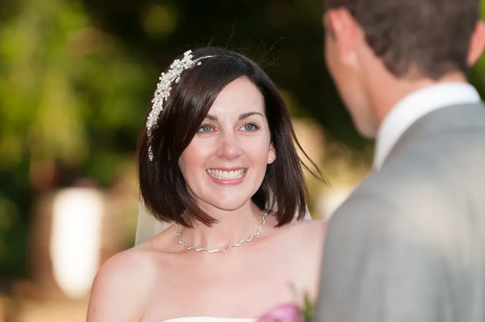 Bride with short dark hair, pearl hair accessory, and silver necklace smiling at groom in gray suit.
