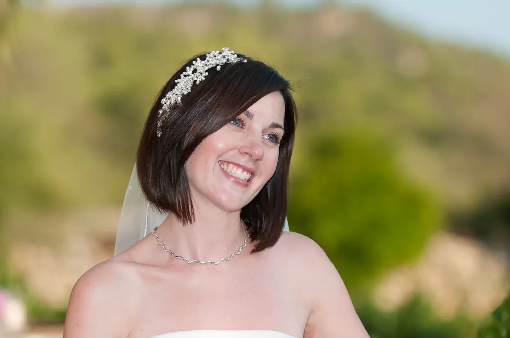 Smiling bride with short dark hair, wearing a jeweled headpiece and necklace against a blurred outdoor background.