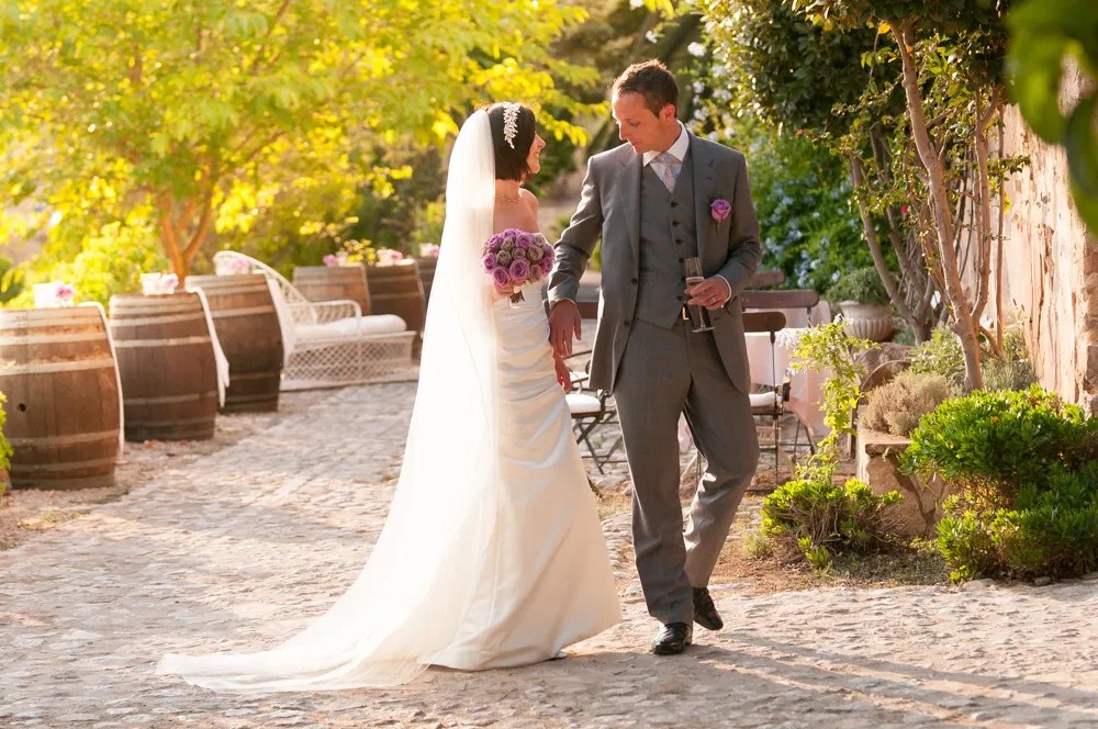 Bride in white gown and veil holding purple bouquet walking hand-in-hand with groom in gray suit outdoors on cobblestone path.