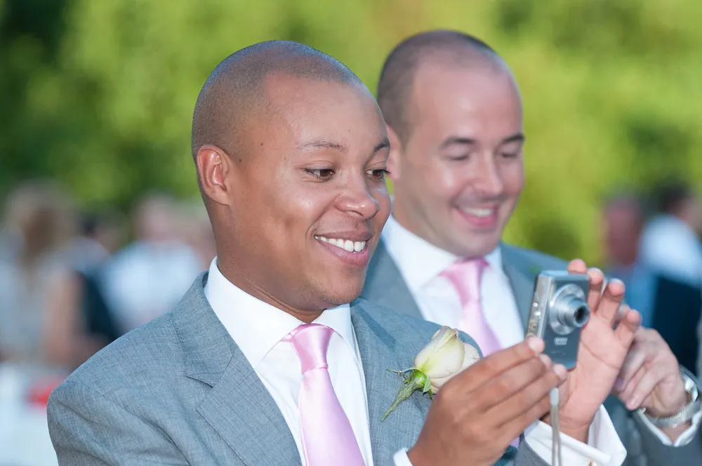 Two men in gray suits with pink ties smiling, one holding and looking at a digital camera with a cream rose boutonniere.