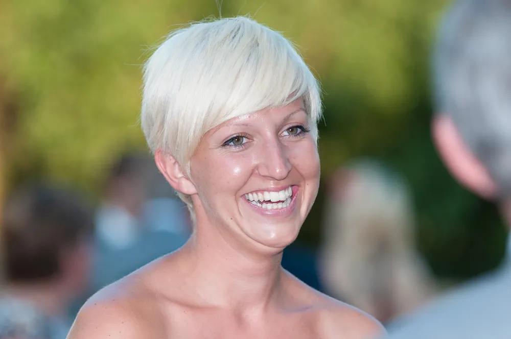 Smiling woman with short platinum blonde hair outdoors, blurred people in background.
