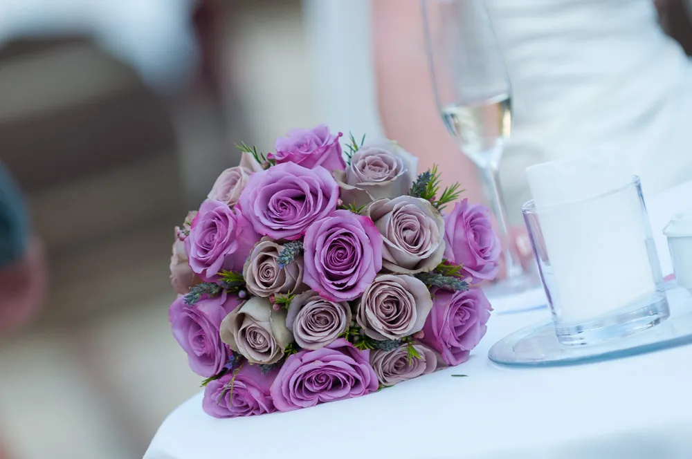 A bouquet of pink and lavender roses placed on a white tablecloth next to a glass with a napkin and a flute glass with champagne.