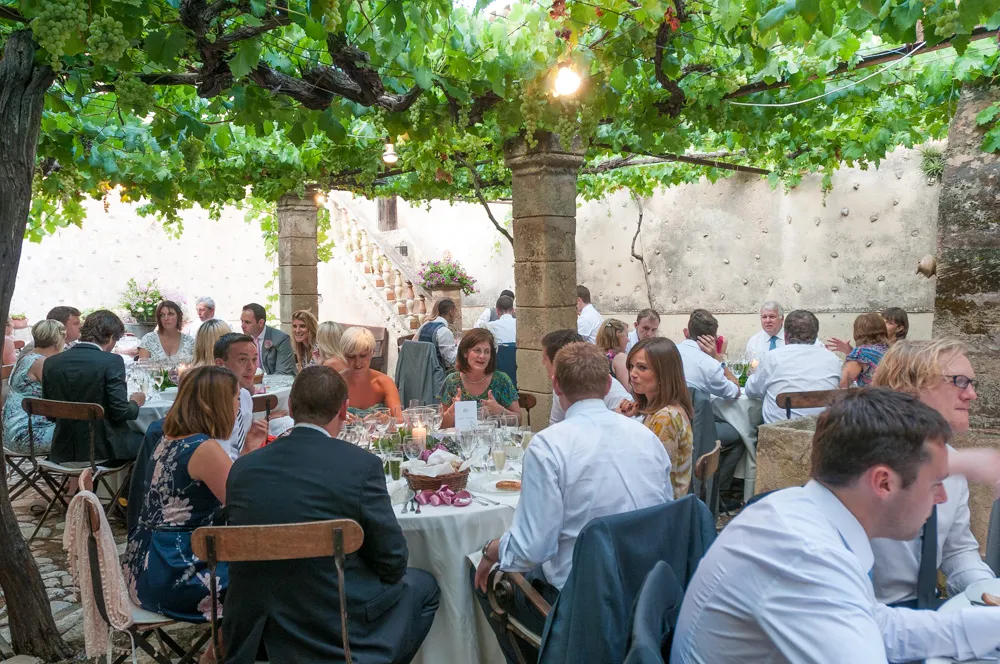 Guests dressed in formal attire dining at round tables under a grapevine-covered pergola in an outdoor courtyard.