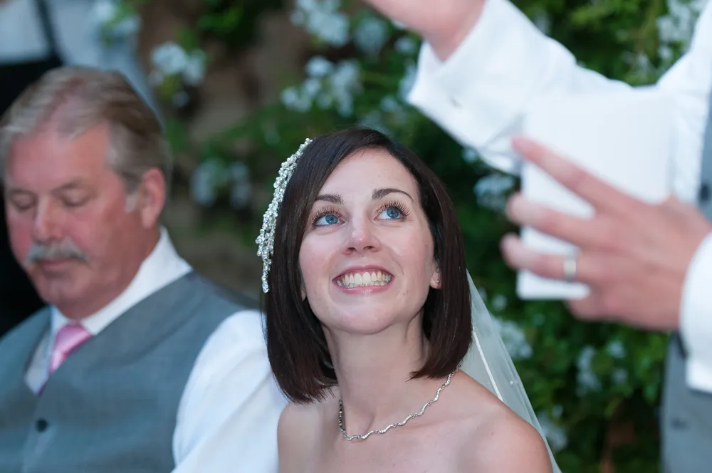 Bride with short brown hair and a jeweled hair accessory smiling and looking up, seated next to an older man in a gray vest and pink tie.