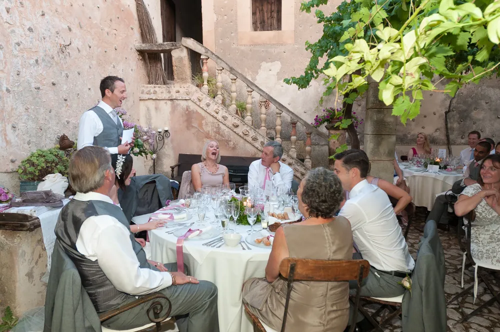 Man in vest and tie giving a speech to guests seated around a round table at an outdoor wedding reception.