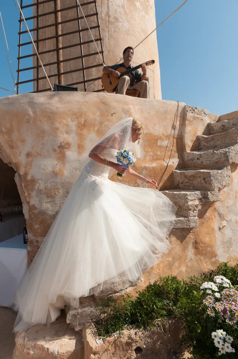 Bride in a white gown and veil holding a blue and white bouquet climbing stone stairs, with a man playing guitar seated above near a windmill.