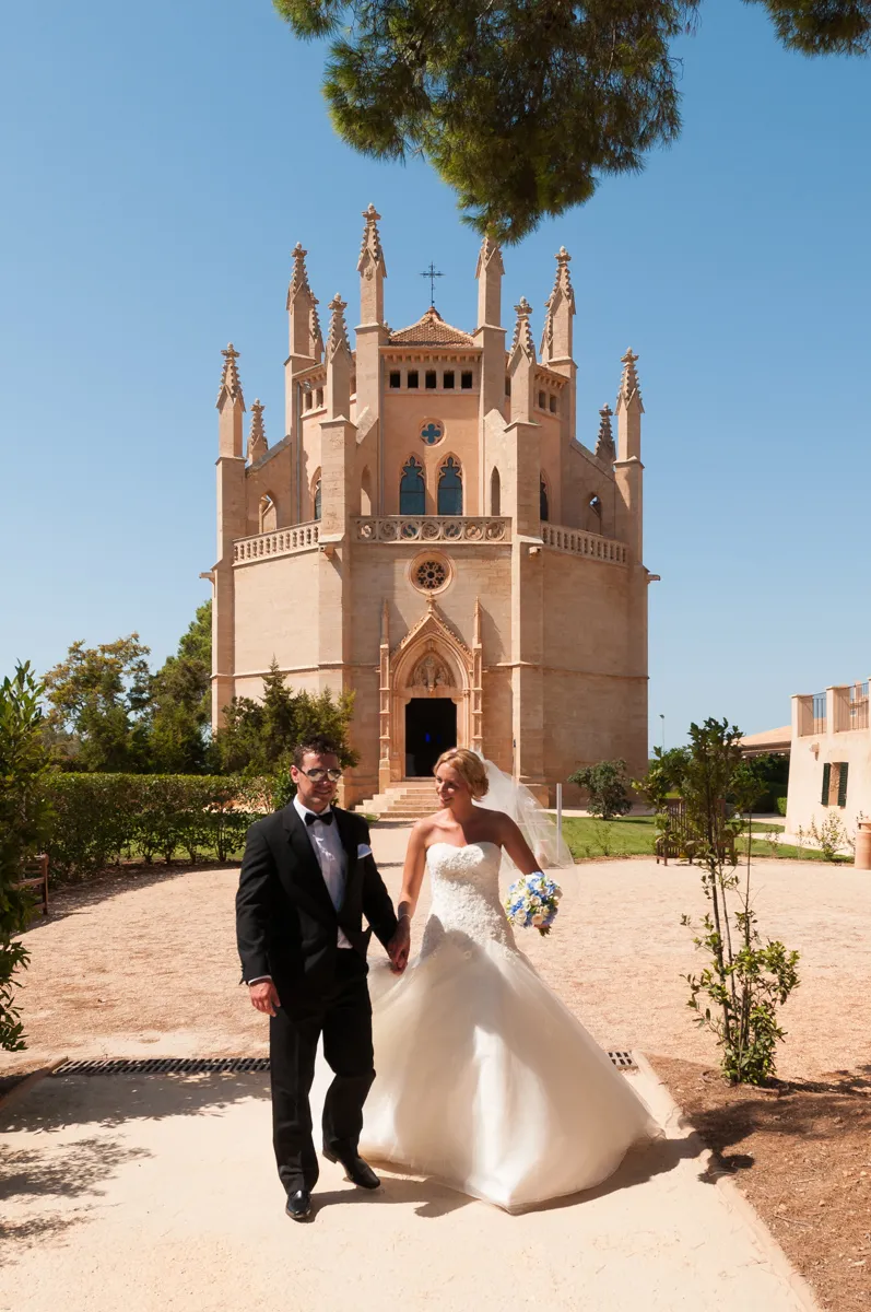 Bride in white wedding dress holding a bouquet and groom in black tuxedo walking hand in hand outside a historic stone church with clear blue sky.