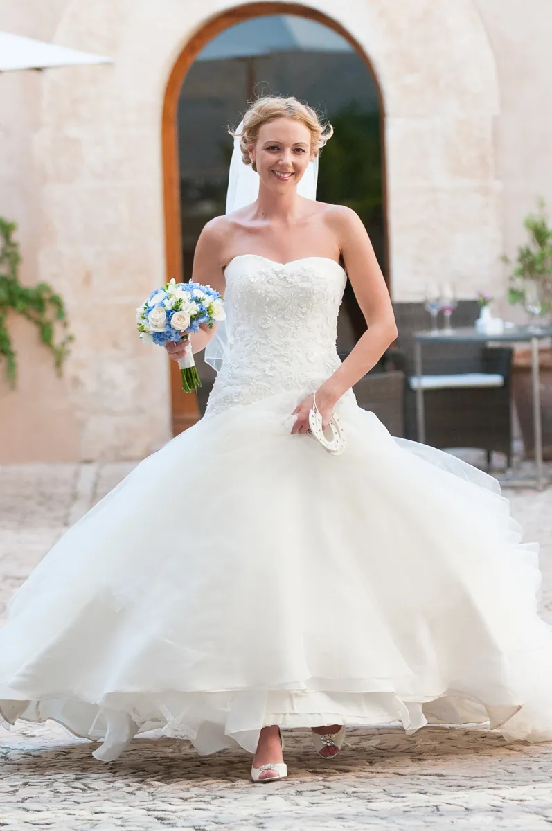 Smiling bride in a strapless white wedding dress holding a bouquet of blue and white flowers walking on a stone pathway.