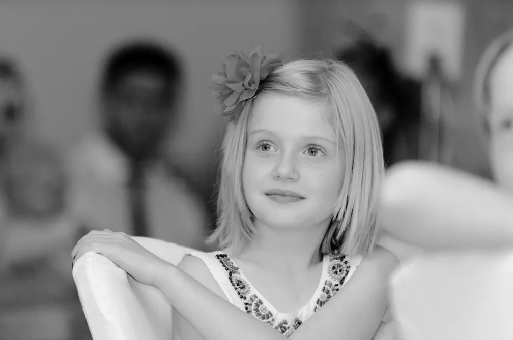 Young girl with a flower in her hair sitting with her arms resting on a chair, looking slightly to the side.