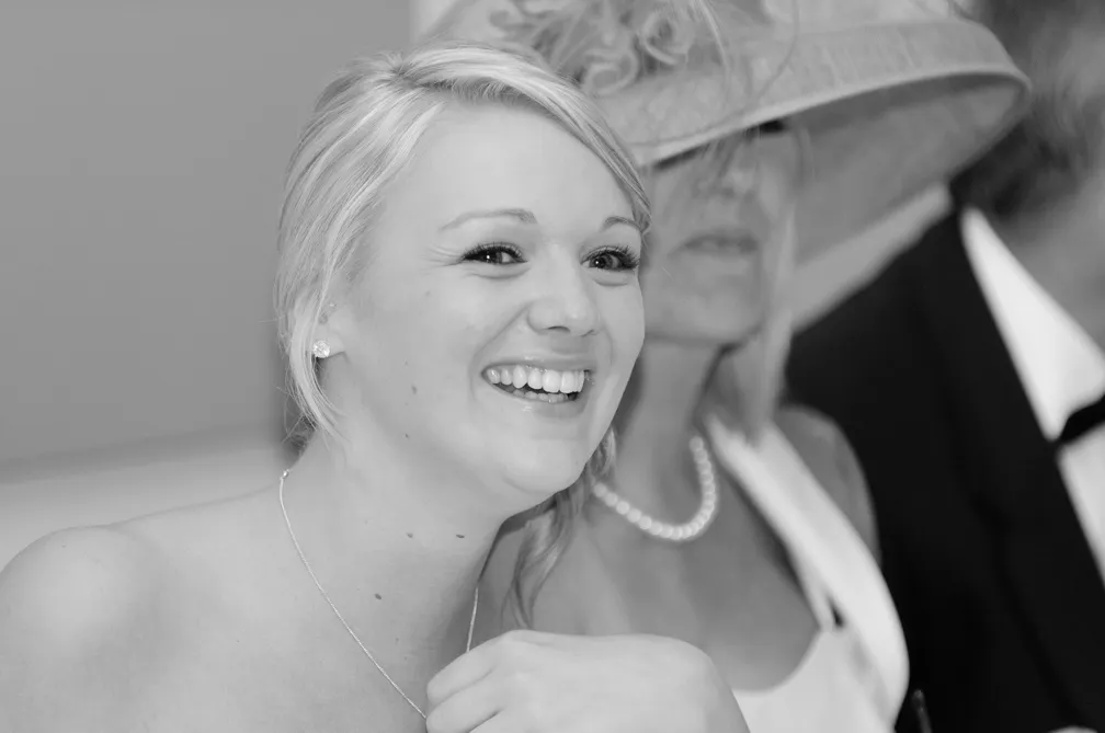 Smiling young woman with blonde hair sitting at a formal event, a woman wearing a large hat and a pearl necklace is blurred in the background.