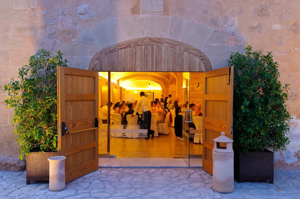 Open wooden doors leading to a warmly lit room where people are seated at tables during an event or dinner.