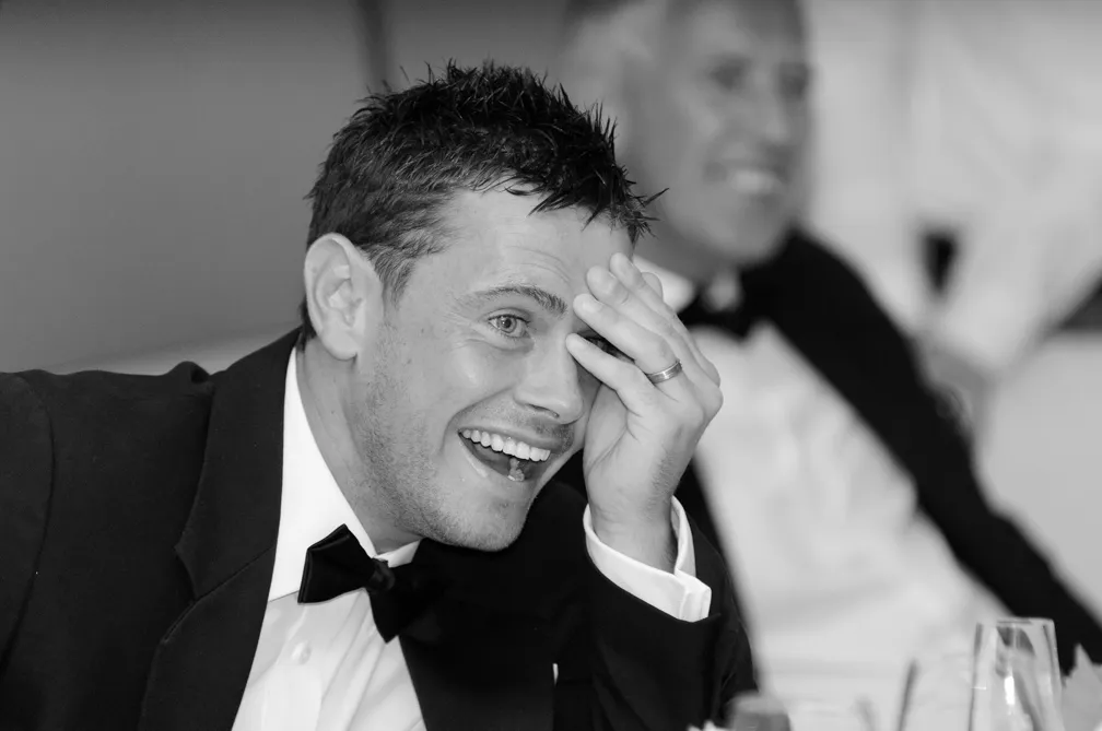 Smiling man in a tuxedo laughing with his hand on his forehead at a formal event.