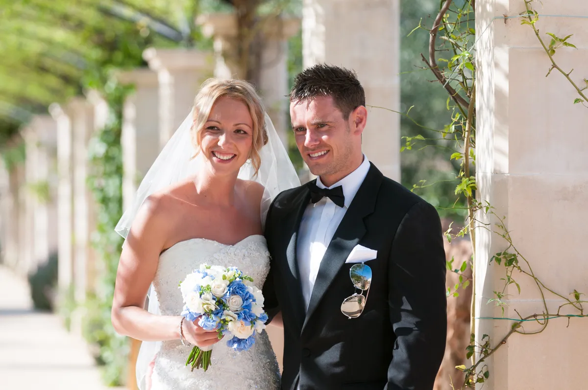 Smiling bride in white wedding dress holding blue and white flower bouquet stands next to groom in black tuxedo with bow tie and sunglasses in pocket.