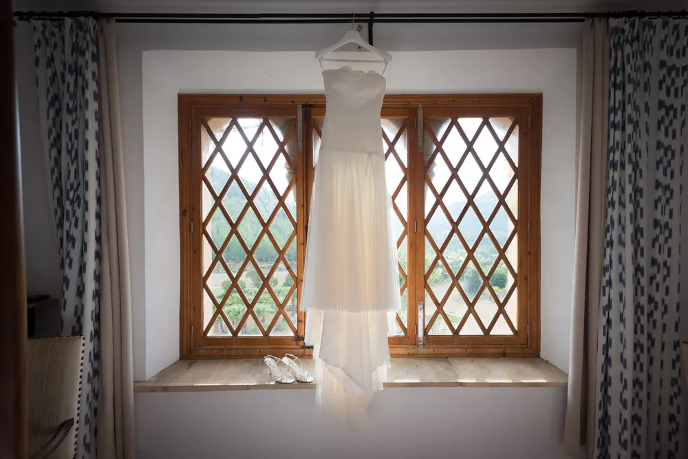 White strapless wedding dress hanging on a hanger in front of a wooden window with diamond-patterned glass panes.
