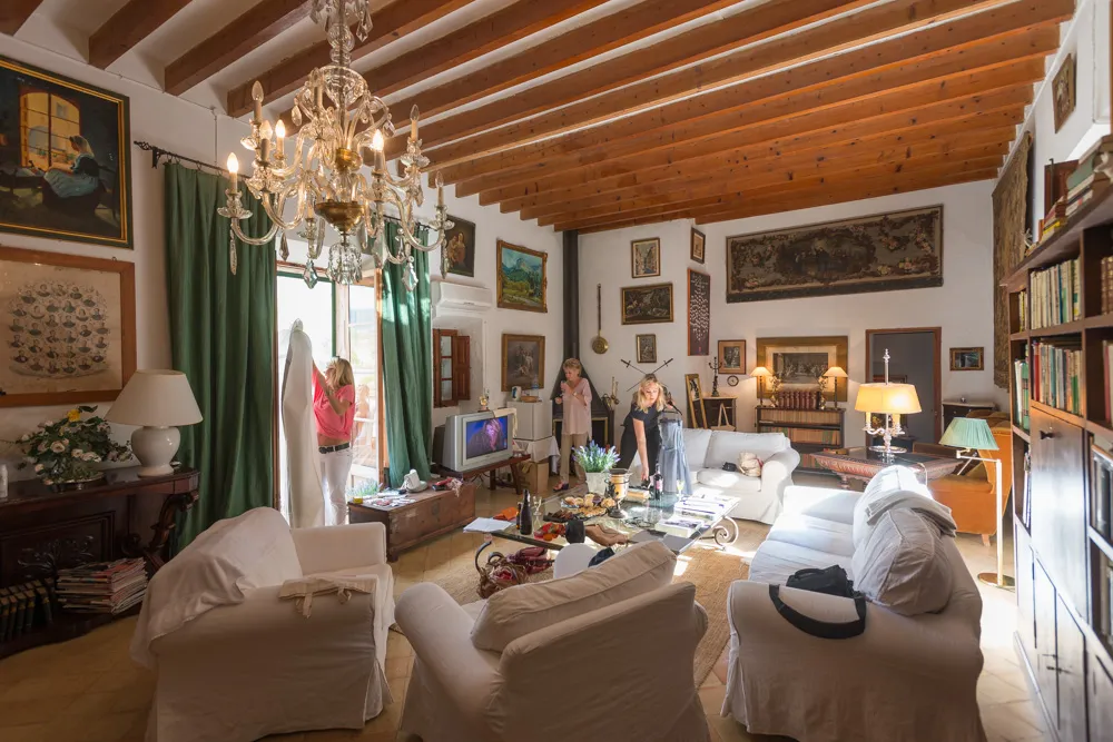 Bright living room with wooden ceiling beams, white furniture, a chandelier, bookshelves, and three people interacting near a glass door with green curtains.