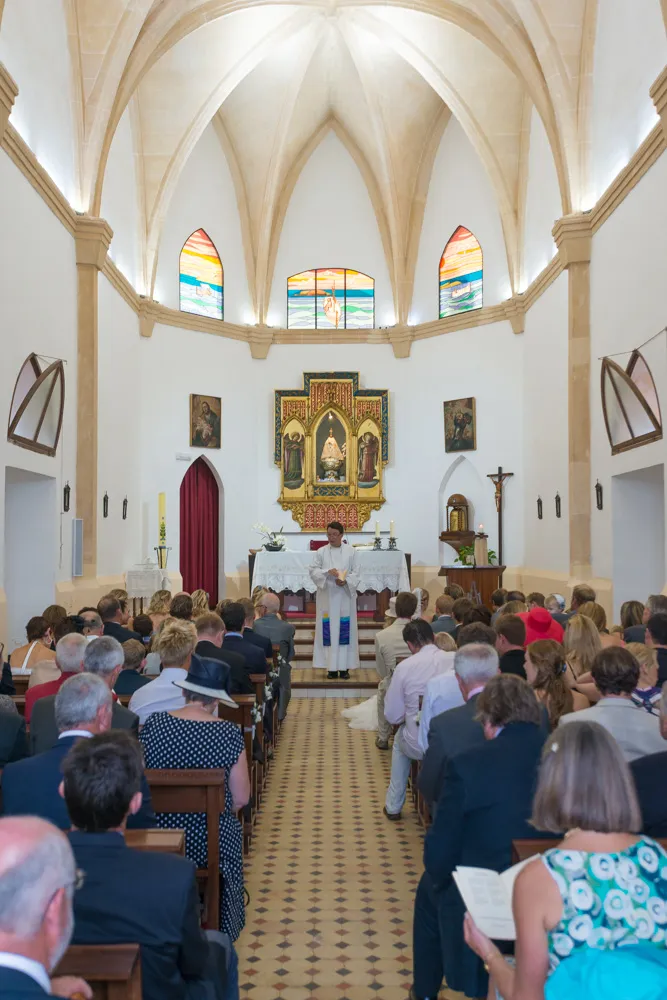 Interior of a church with a priest standing at the altar reading during a wedding ceremony, with guests seated in wooden pews.