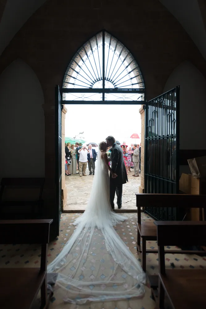 Bride and groom kissing while holding hands at a church doorway with guests standing outside under umbrellas.