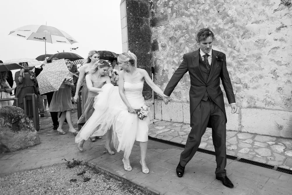 Black and white photo of a bride and groom holding hands walking outside with bridesmaids and guests holding umbrellas in the rain.