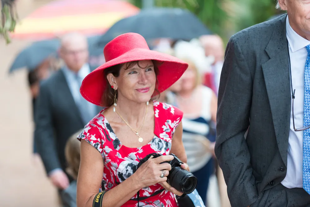 Woman in a red floral dress and wide-brimmed red hat holding a camera at an outdoor event.