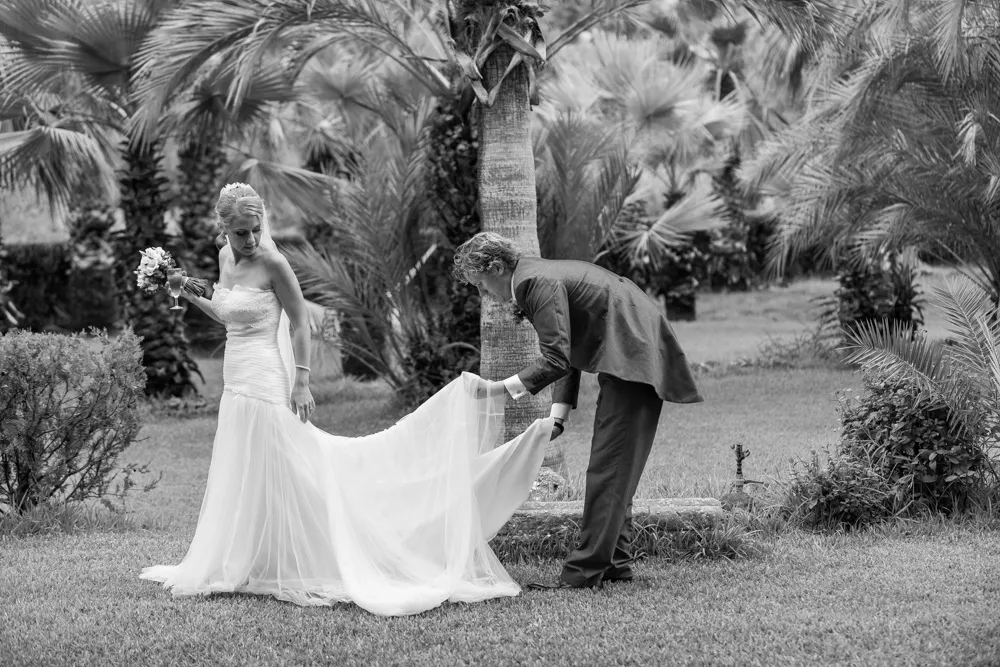 Bride in a strapless wedding gown holding a bouquet and glass while groom adjusts the train in a garden with palm trees.