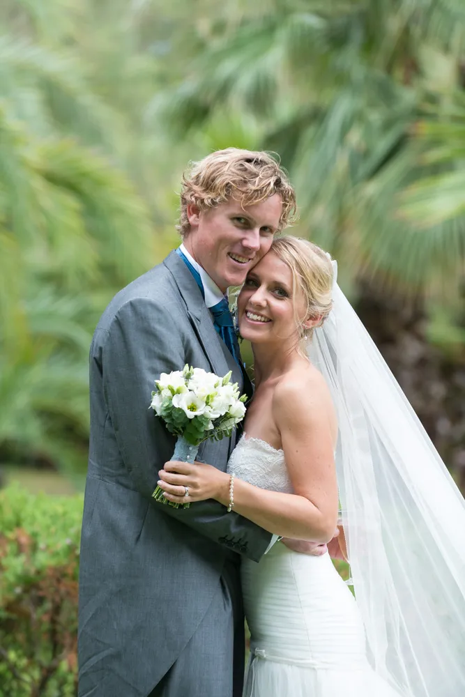 Bride in a white strapless gown and veil hugging groom in a gray suit holding a white flower bouquet outdoors with green foliage background.