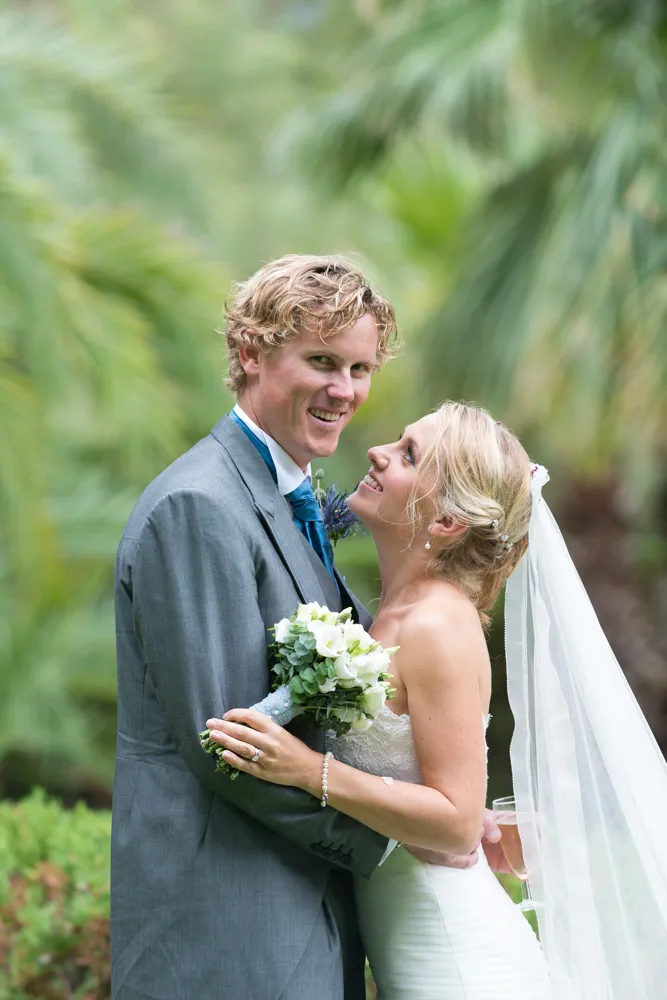 Bride and groom embracing outdoors, the bride holding a bouquet and wearing a veil, the groom in a gray suit with a blue tie.