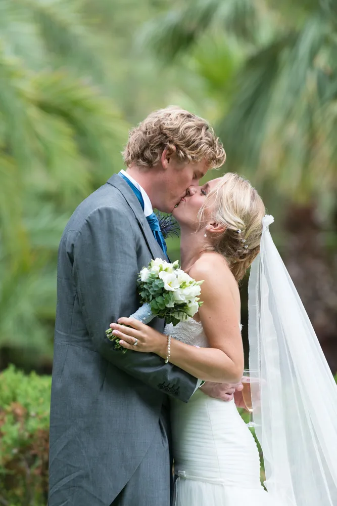 Bride and groom in wedding attire kissing outdoors with greenery background, bride holding bouquet and wearing veil.