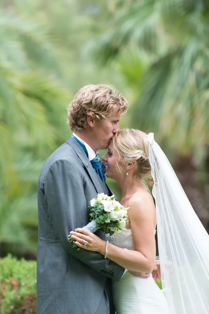 Groom kissing bride's forehead as they embrace outdoors, bride holding white flower bouquet and wearing veil.