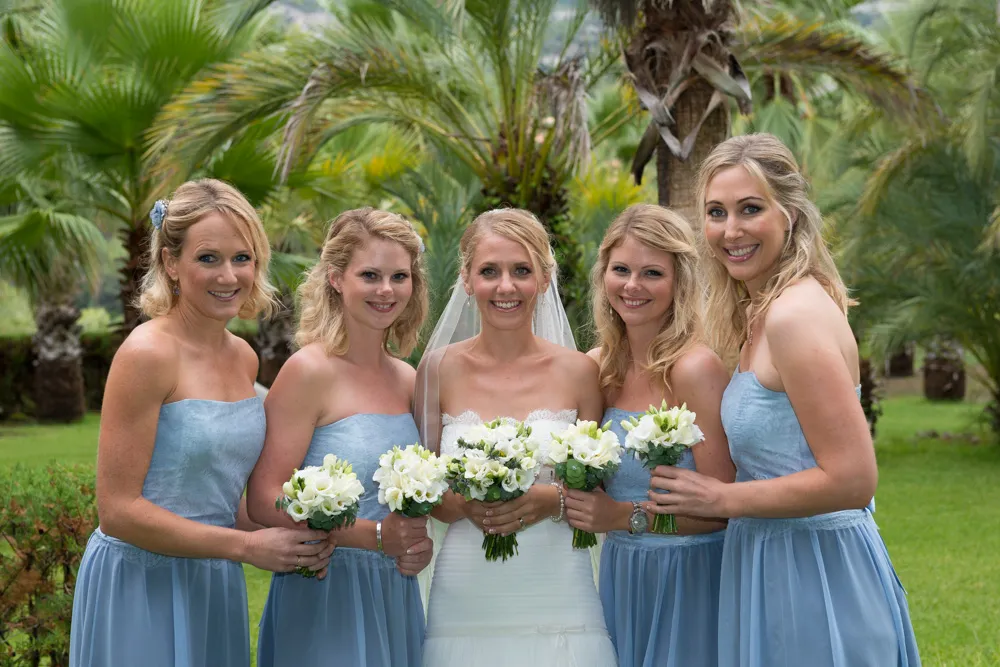 Bride in white wedding dress with veil stands smiling outdoors with four bridesmaids wearing light blue strapless dresses, all holding white floral bouquets.
