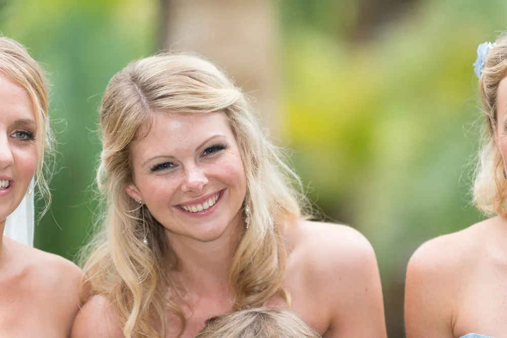 Smiling woman with long blond hair wearing pearl earrings outdoors with blurred green background.