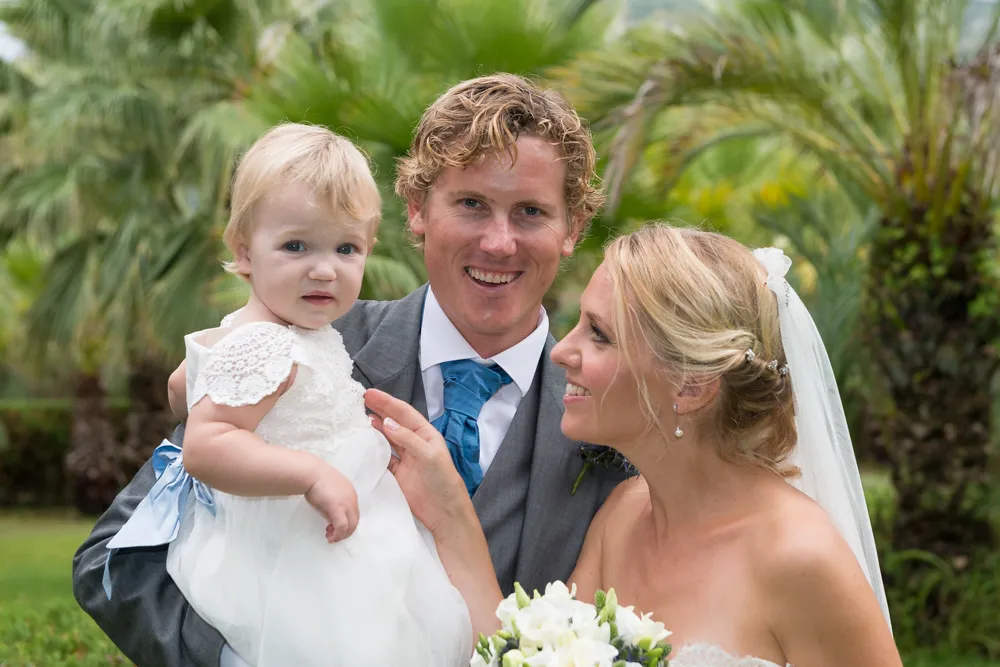 Smiling bride and groom outdoors holding a young child dressed in white with palm trees in the background.