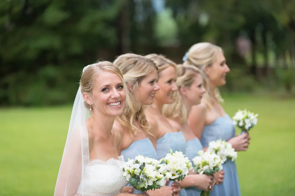 Bride smiling and standing outdoors with four bridesmaids in light blue dresses holding white flower bouquets.