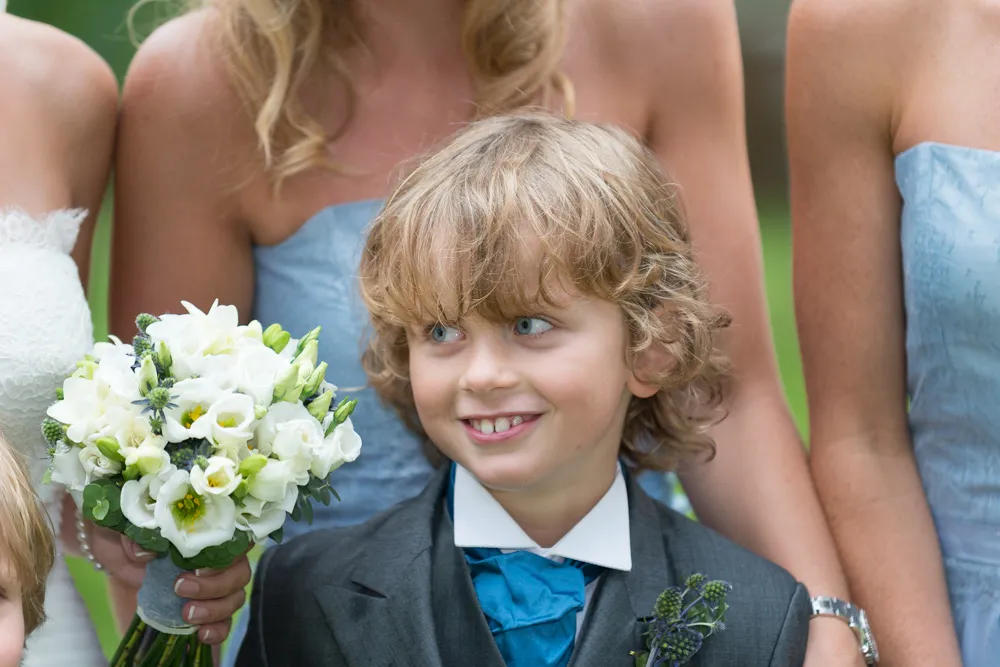 Young boy in a suit with a blue bow tie smiling, surrounded by women in blue dresses holding a white flower bouquet.