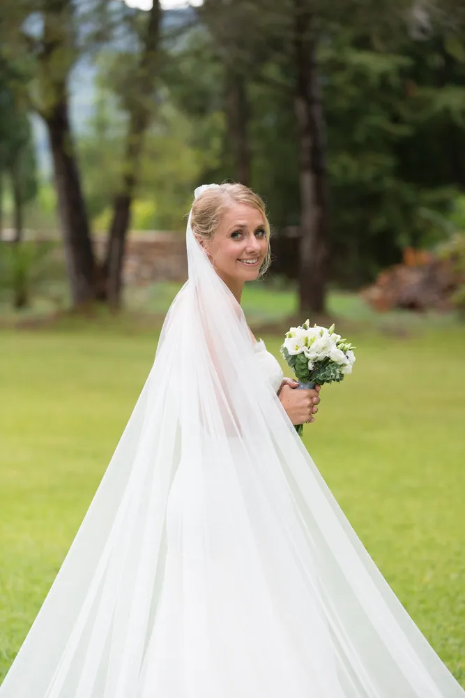 Smiling bride in a white wedding dress and long veil holding a bouquet of white flowers outdoors on a grassy area.