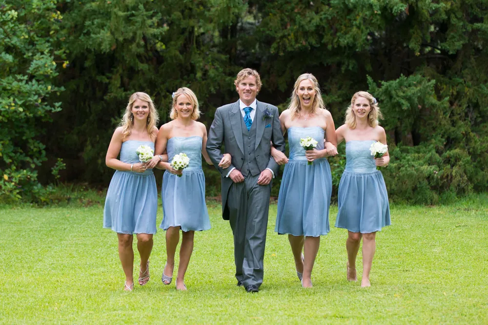 Man in gray suit walking arm-in-arm outdoors with four bridesmaids in light blue dresses holding small white flower bouquets.