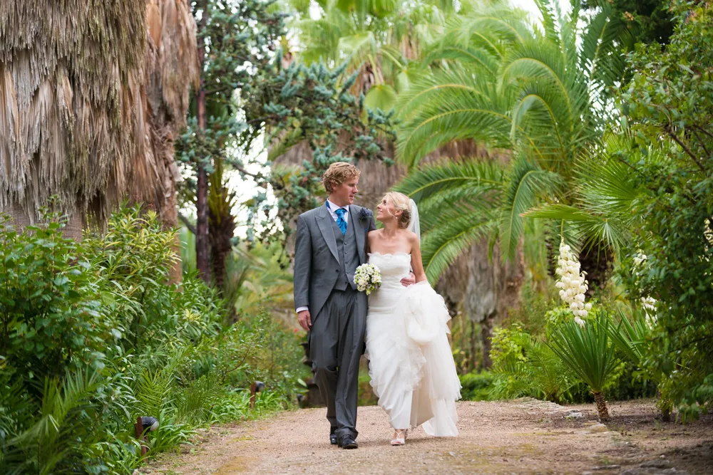 Bride in white wedding dress and groom in gray suit walking arm in arm on a garden path surrounded by lush greenery and palm trees.