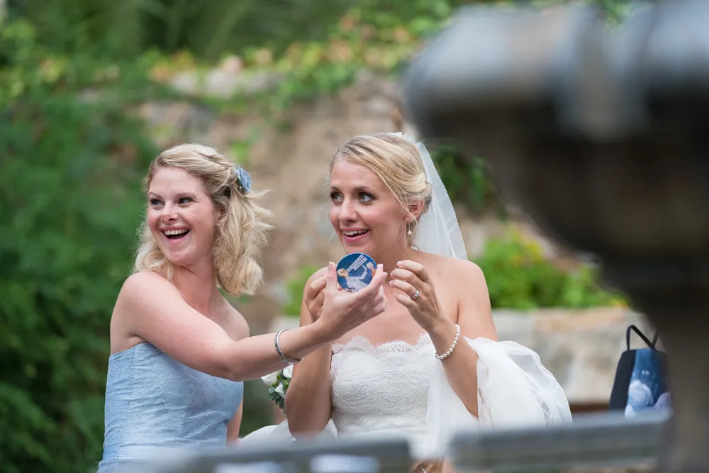Bride in lace wedding dress and veil shares a joyful moment holding a small blue round object with a bridesmaid in a light blue strapless dress outdoors.