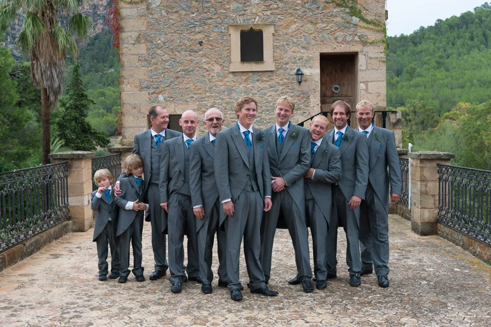 Group of men and two boys in matching gray suits with blue ties standing and smiling outdoors in front of a stone building.