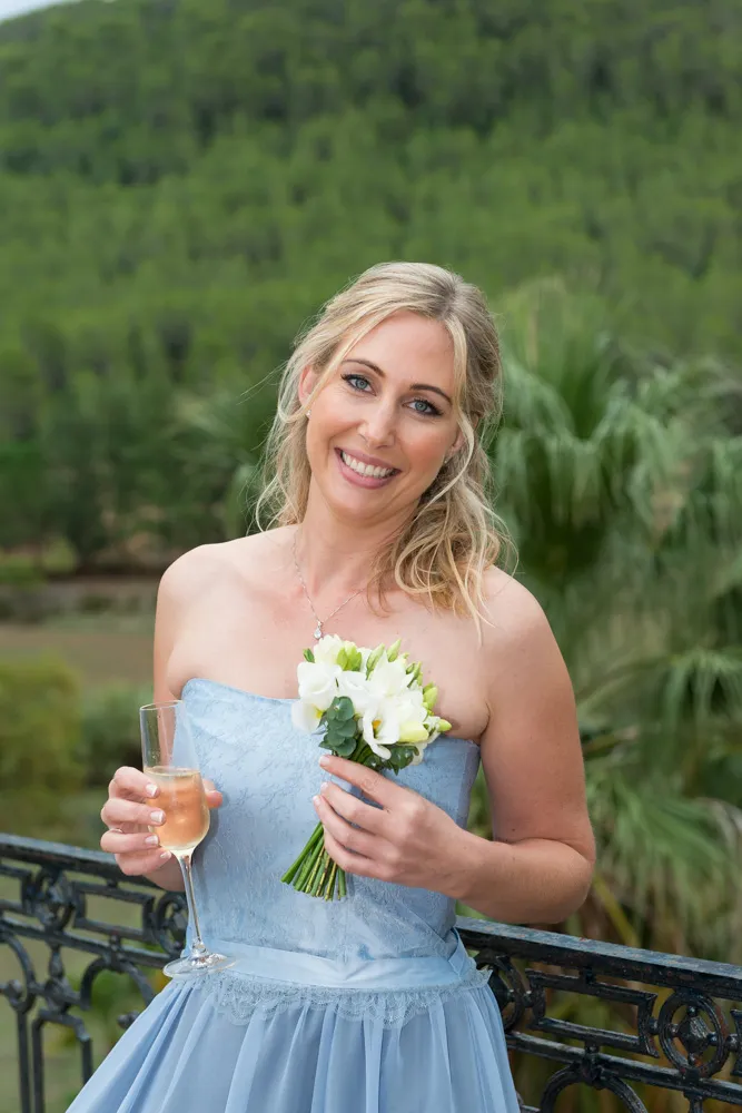 Smiling woman in a light blue strapless dress holding a small white flower bouquet and a glass of champagne outdoors with greenery in the background.