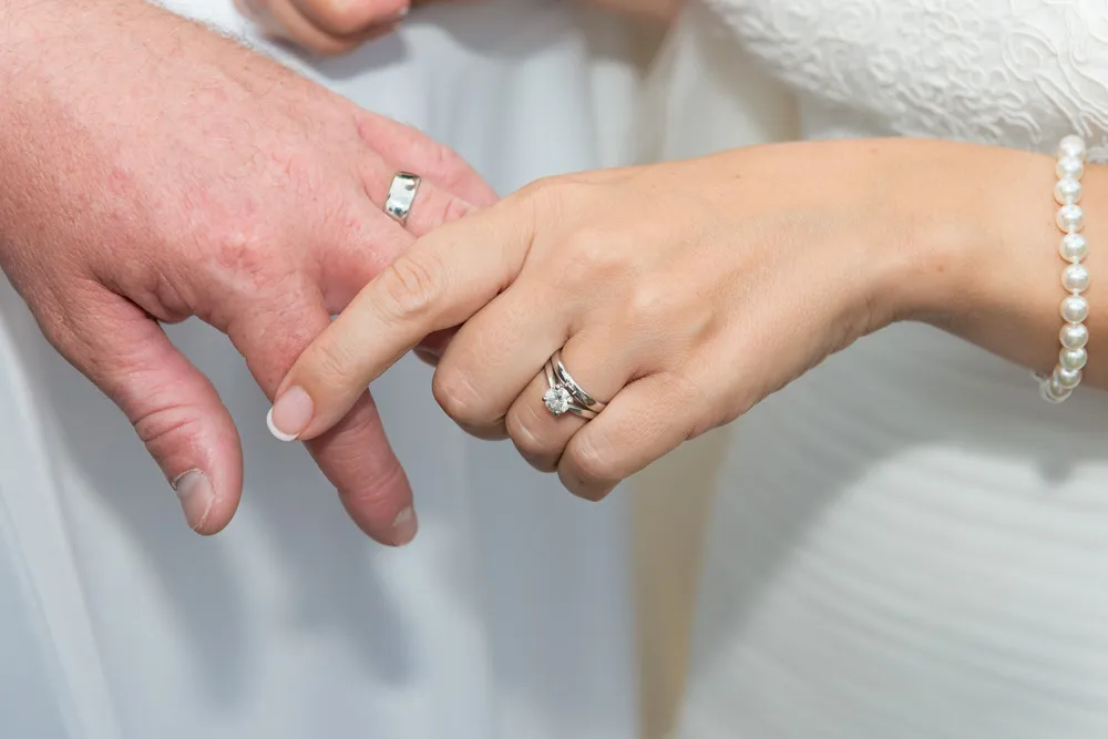 Close-up of a bride and groom's hands showing wedding rings, with the bride wearing a pearl bracelet and white dress.