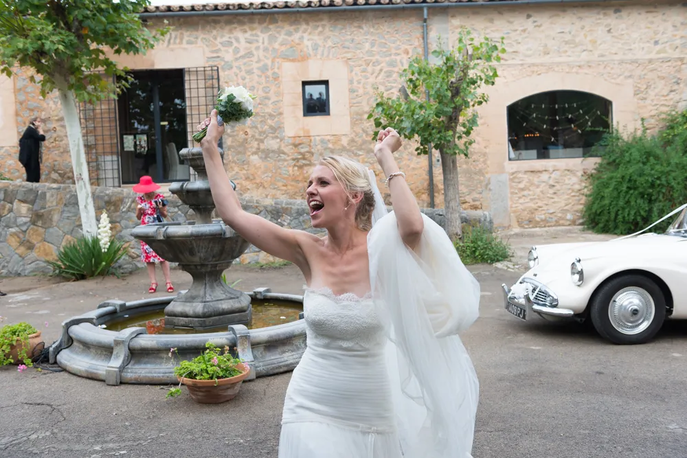 Joyful bride in white wedding dress holding bouquet with arms raised near stone fountain and vintage white car.