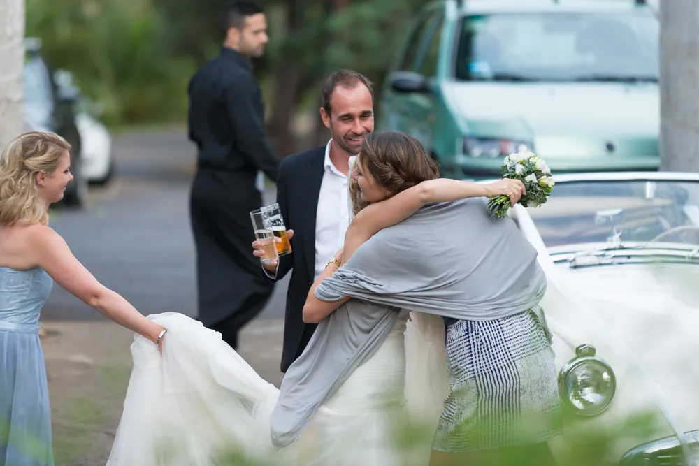 Bride hugging a woman holding a bouquet near a white vintage car, with a man holding two glasses and a bridesmaid adjusting the dress.
