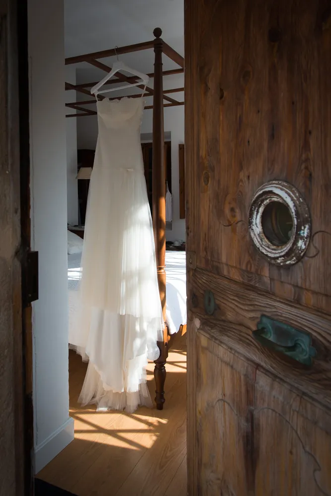 White wedding dress hanging on a wooden four-poster bed in a sunlit room with a partially open wooden door in the foreground.