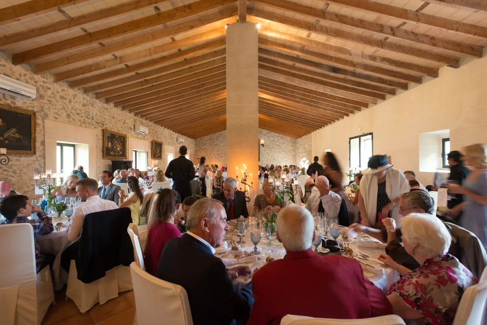 Large group of people seated at round tables inside a rustic room with wooden beams and stone walls, enjoying a formal event or celebration.