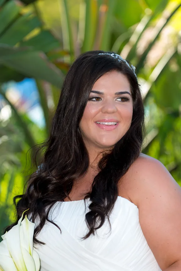 Smiling woman with long dark hair wearing a white strapless dress and holding white flowers, with green leaves in the background.