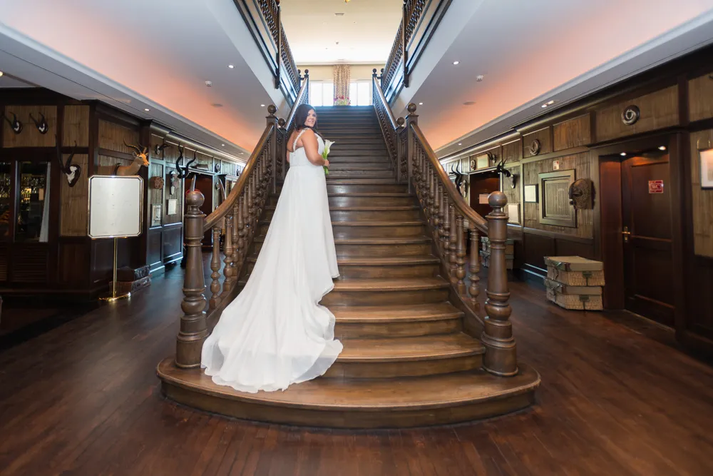 Bride in a white wedding gown holding flowers standing on wooden stairway in a rustic interior with wooden walls and mounted animal heads.
