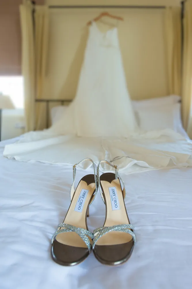 Pair of glittery silver Jimmy Choo high-heeled sandals placed on a white bed with a white wedding dress hanging in the background.