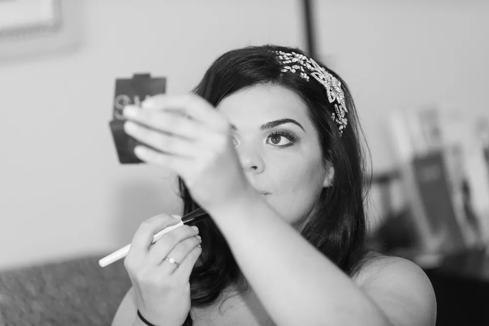Woman with jeweled hairpiece applying makeup using a small mirror.